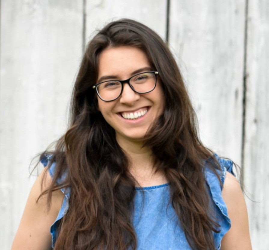 Woman with glasses in a blue top smiling and standing in front of a greyish white background