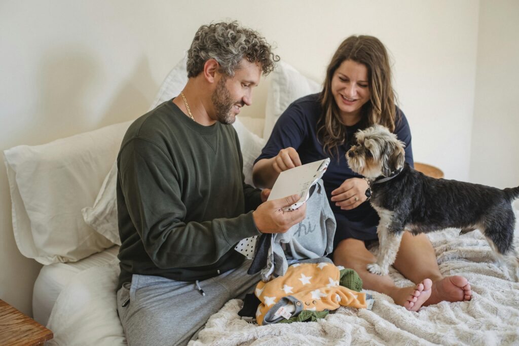A couple joyfully preparing baby clothes in a cozy bedroom with their attentive dog to put in a baby keepsake box.