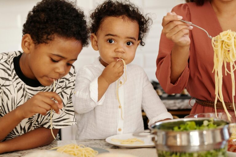 Two young children eating pasta at home with family, enjoying a fun meal together.