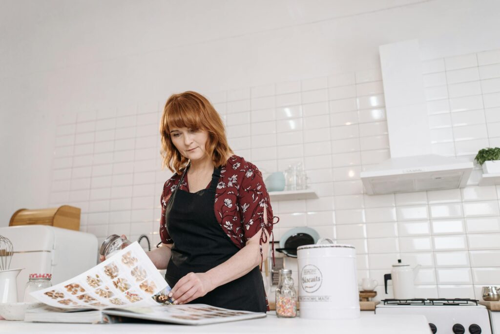 Looking at cookbook Photo by Pavel Danilyuk A woman in the kitchen examining a recipe book, to improve meal planning skills