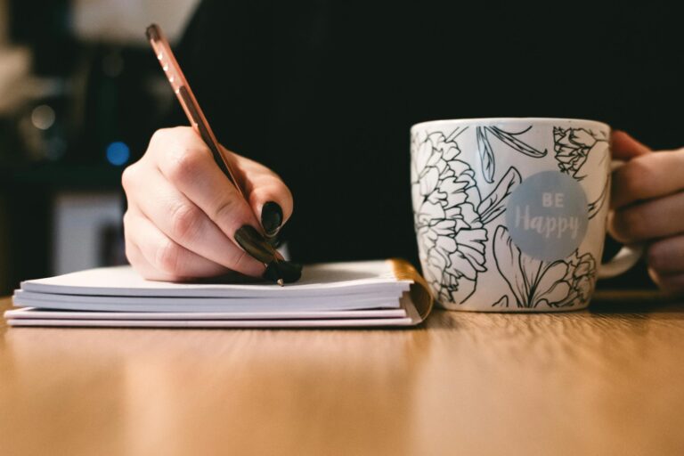 Person writing a birth story in a notebook with a floral ceramic mug on a wooden desk.