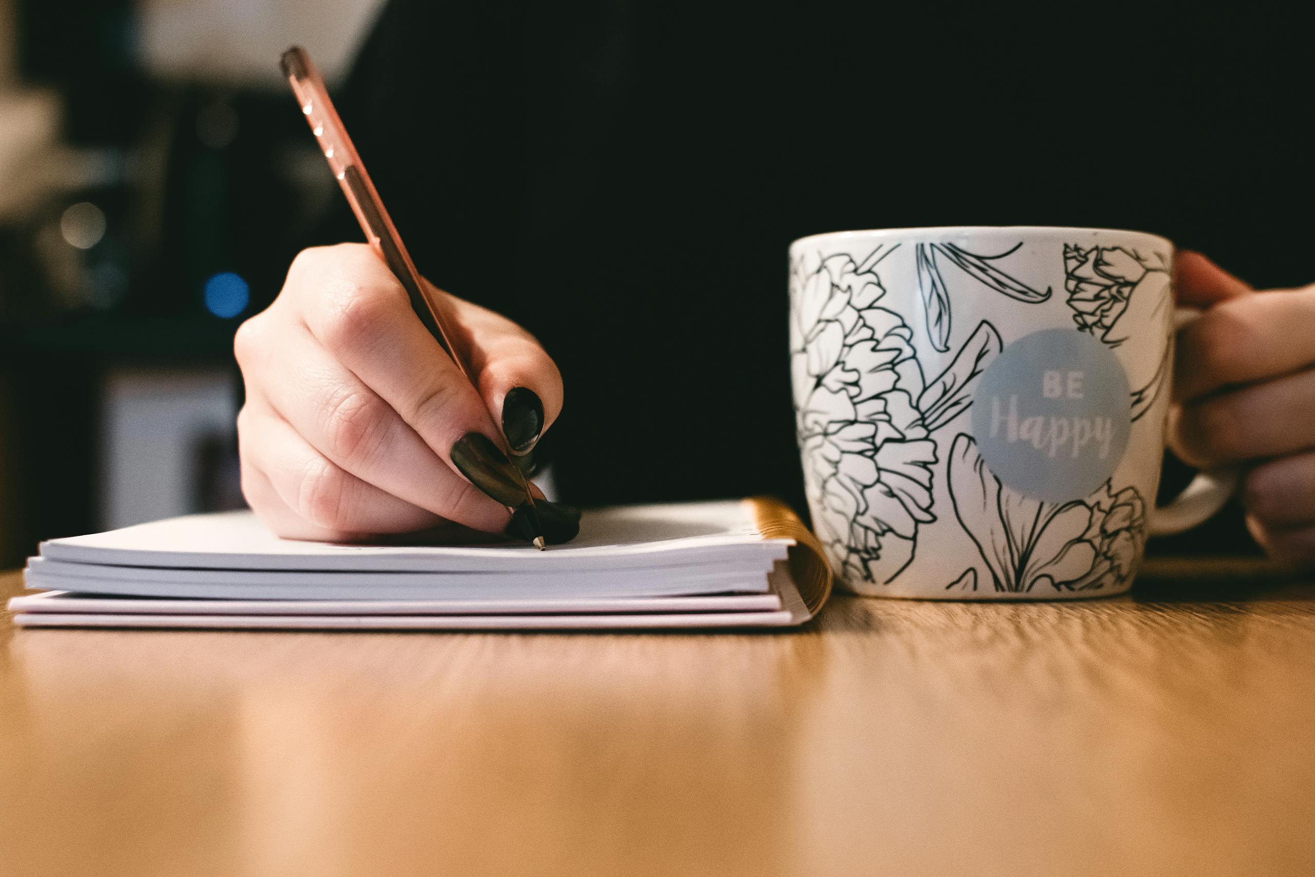 Person writing a birth story in a notebook with a floral ceramic mug on a wooden desk.